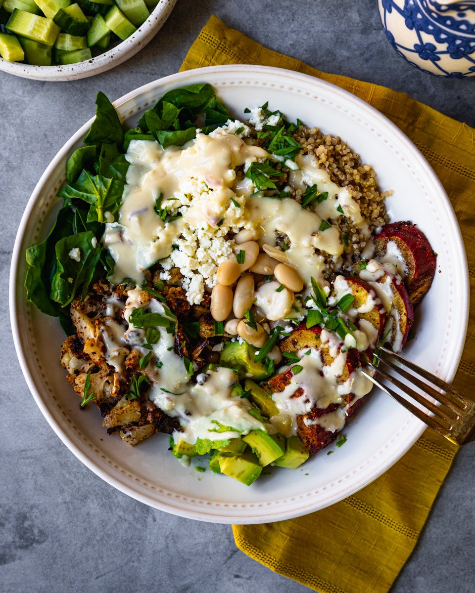 Top down view of a completed quinoa bowl with beans, quinoa, cucumbers, spinach, sweet potatoes, cauliflower, avocado, feta and tahini sauce